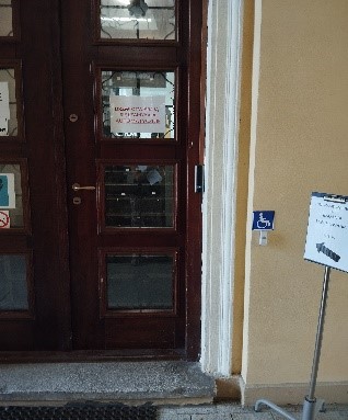 Wejście dla klientów urzędu w Warszawie. Przed drzwiami tabliczka z napisem do badania i cechowania ze strzałką w kierunku drzwi. Na ścianie piktogram oznaczający niepełnosprawność, pod nim  przycisk dzwonka.