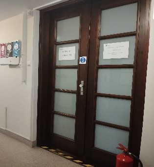 Fragment korytarza z podwójnymi brązowymi drzwiami do pokoju obsługi. Na ścianie wiszą kolorowe obrazki przy drzwiach po prawej stronie na szarej podłodze stoi czerwona gaśnica przeciwpożarowa.